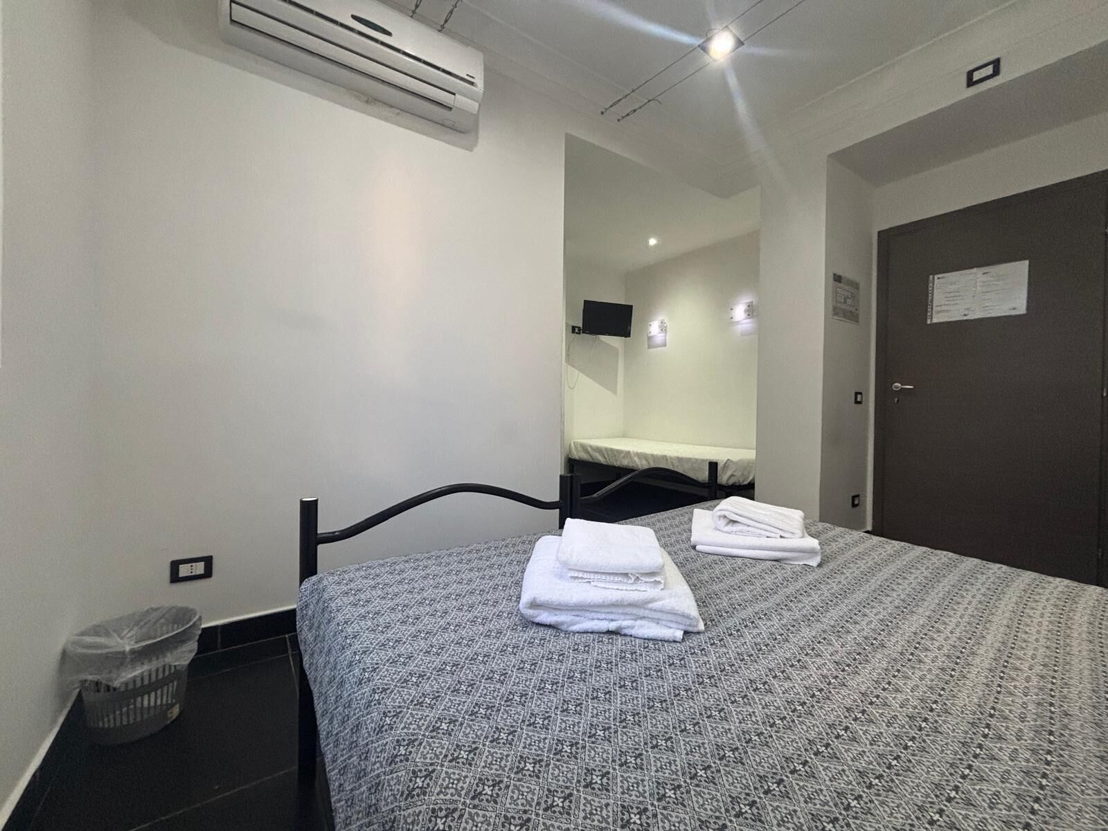 Habitación de hotel con cama, aire acondicionado de pared, un cesto de basura pequeño y una puerta con un cartel.