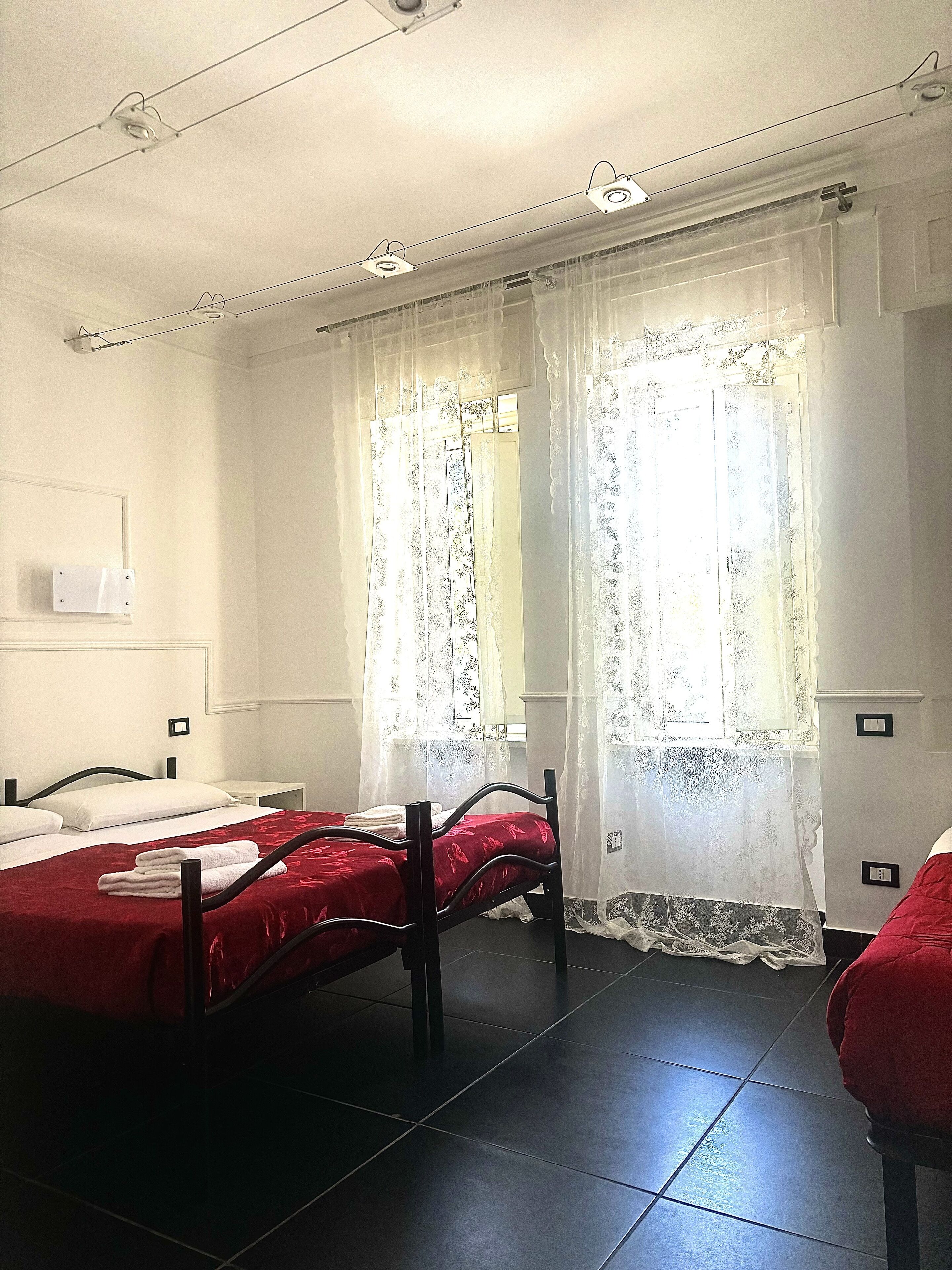 Habitación de hotel con dos camas individuales, una mesita de noche pequeña, una ventana con cortinas de gasa y suelo de baldosas.
