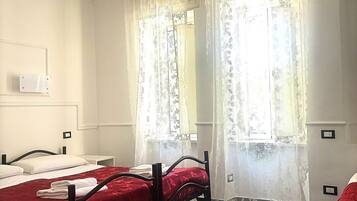 Une chambre d’hôtel avec deux lits simples, une petite table de chevet, une fenêtre avec des rideaux légers et un sol carrelé.