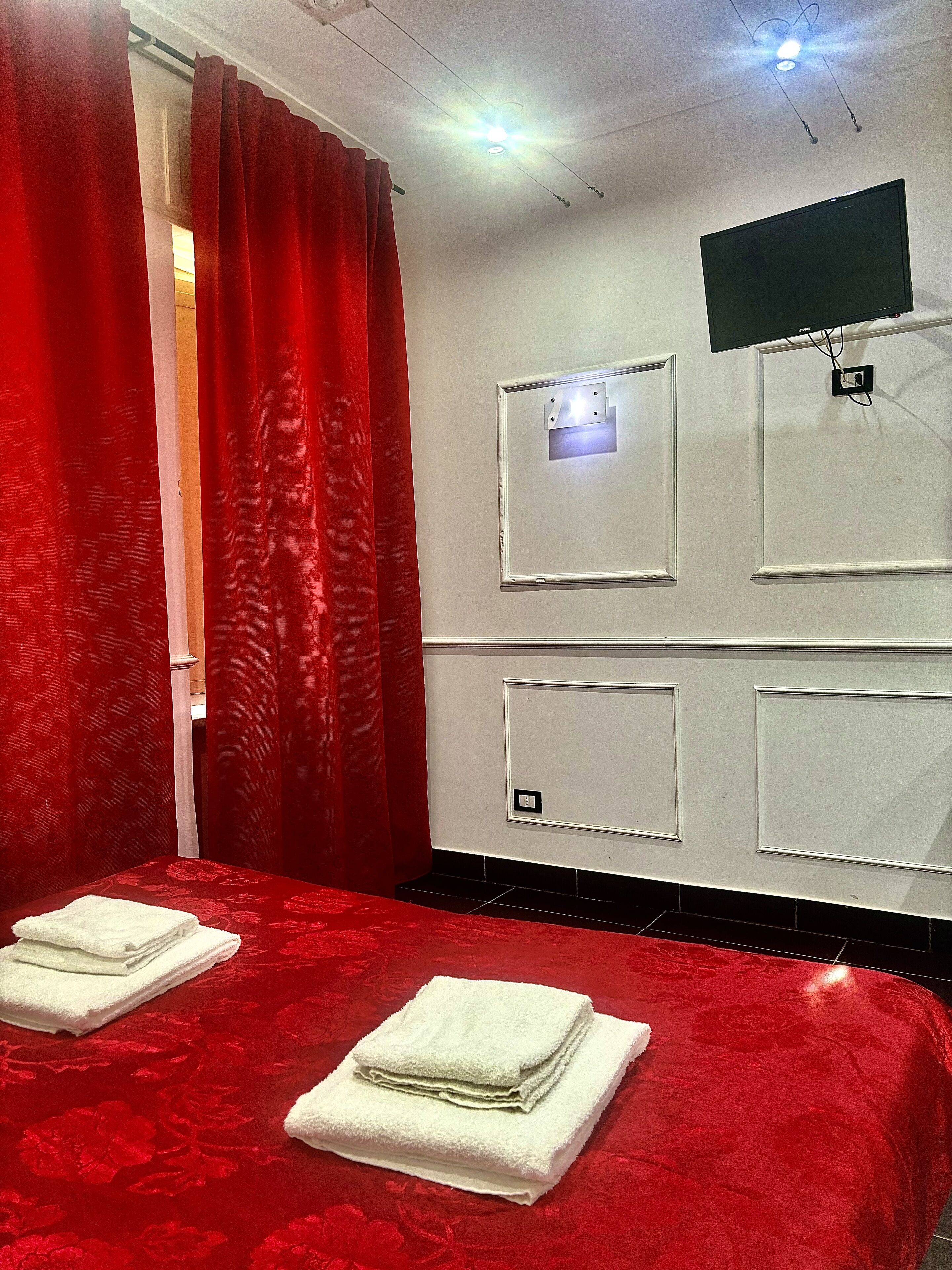 Camera d'albergo con copripiumino rosso, due asciugamani bianchi, una TV a schermo piatto montata a parete e una ventola a soffitto.