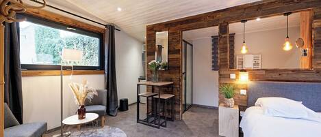 Un dormitorio moderno con cama, mesita de noche y un espejo grande. Hay una ventana con cortinas, una pequeña mesa con un florero y una silla.