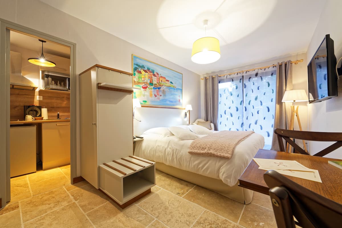 Habitación de hotel pequeña con cama, escritorio, silla, televisor, un cuadro y kitchenette.
