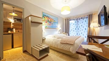 Een kleine hotelkamer met een bed, bureau, stoel, televisie, schilderij en kitchenette.