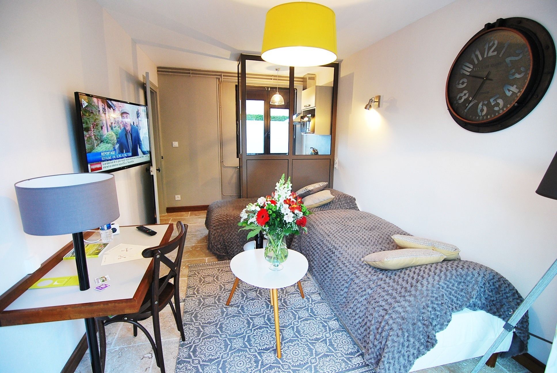 A compact hotel room with a bed, a desk with a chair, a TV, a mirror, and a clock on the wall.