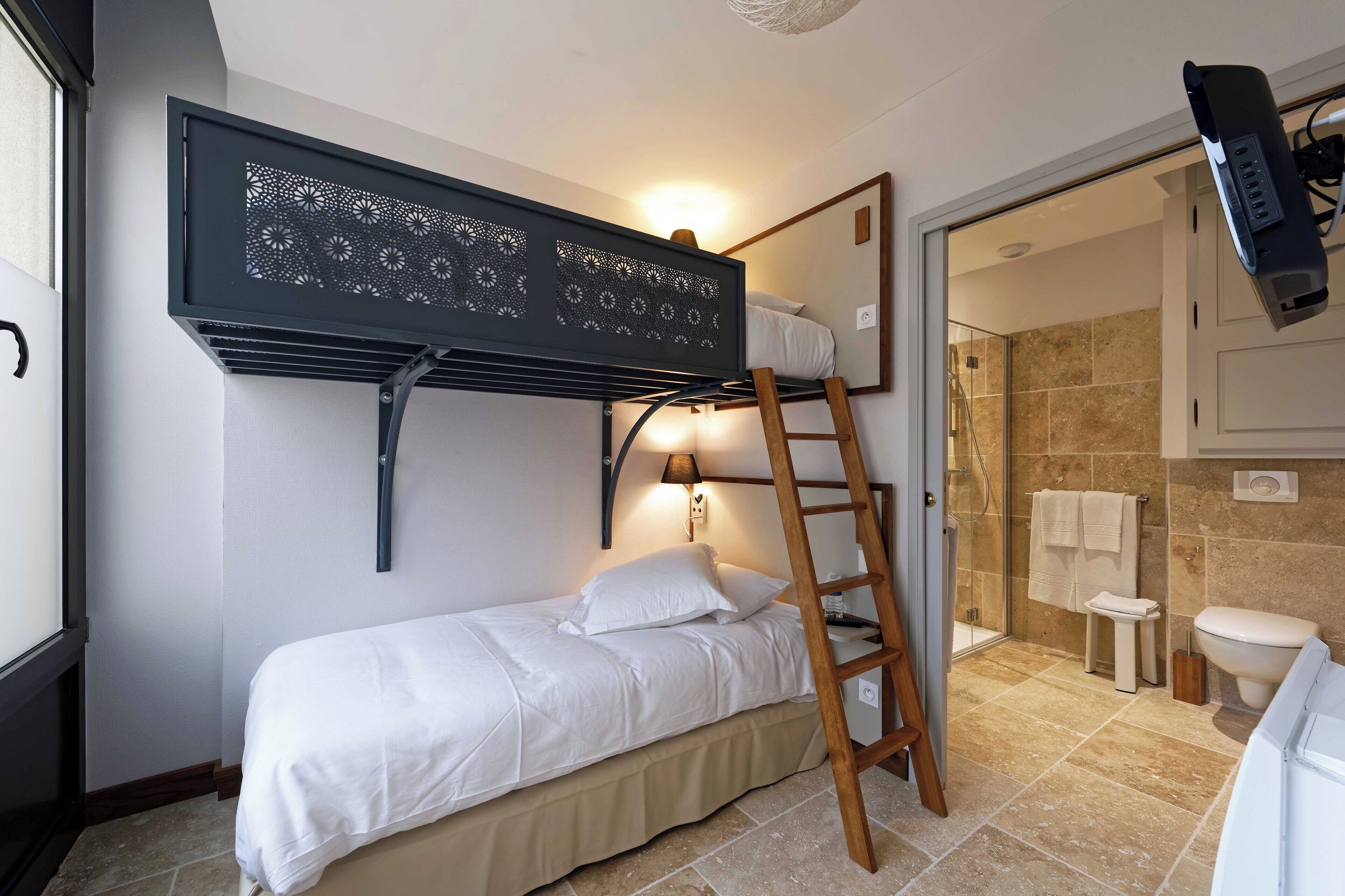 A compact bedroom with a bunk bed, a ladder, a bedside table, a wall-mounted TV, and a bathroom with a shower and toilet.