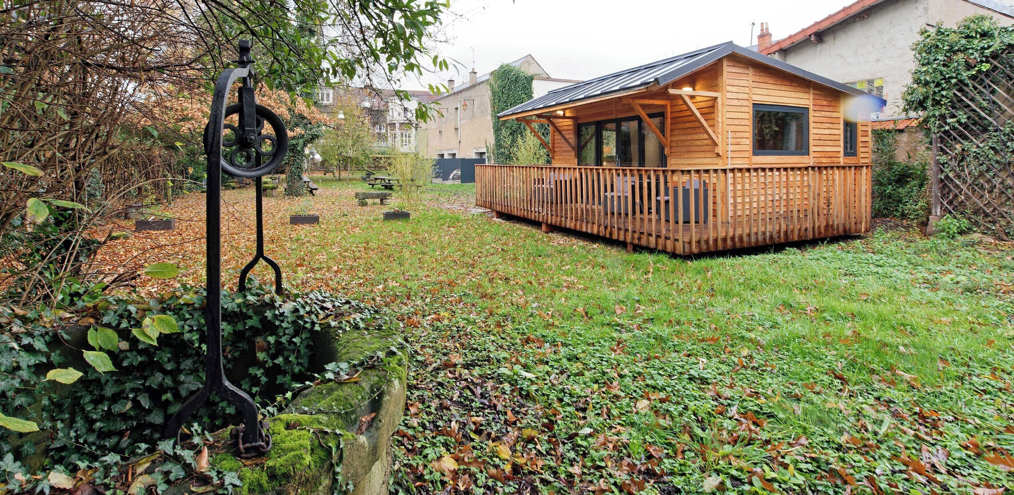 Une cabane en bois dotée d’une terrasse, entourée d’un jardin où se trouvent un banc en métal et un arbre.