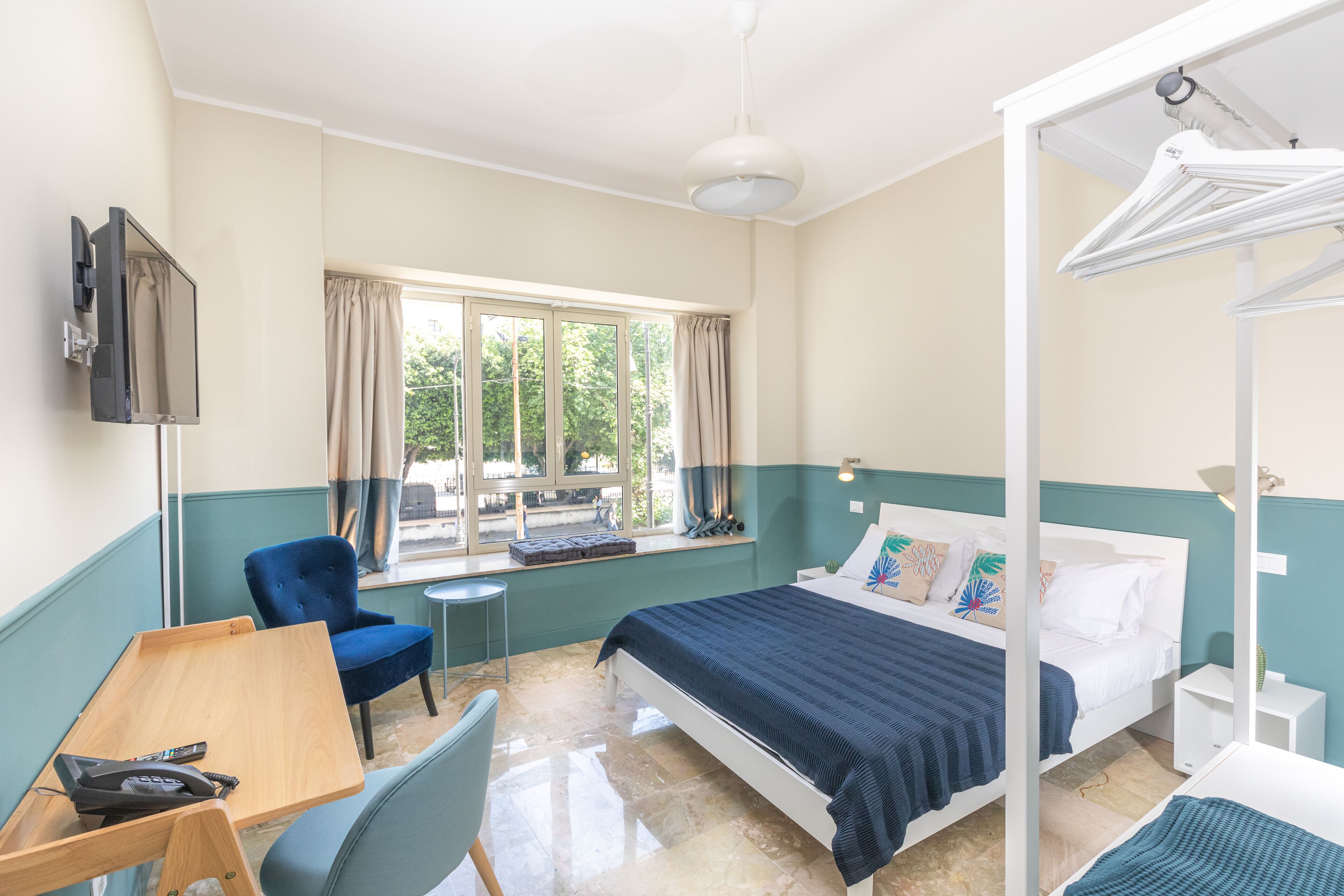 A bedroom with a bed, a desk, a chair, a window, and a hanging light fixture.