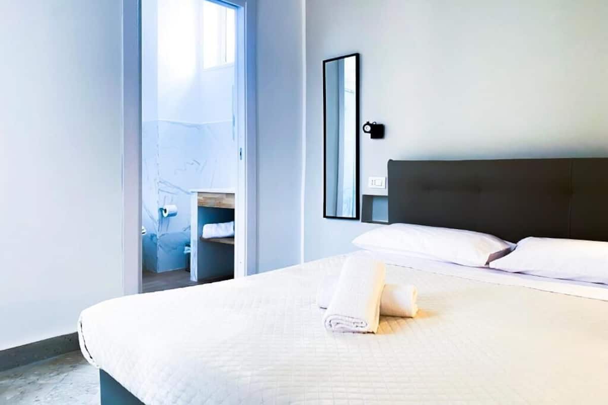 A hotel room with a bed, a bedside table, a mirror, and a door leading to a bathroom.