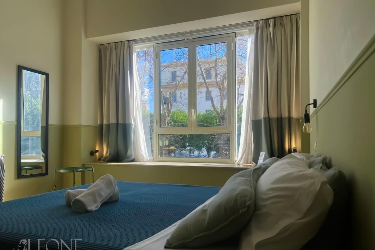 A hotel room with a bed, a window with curtains, a nightstand, and a mirror.