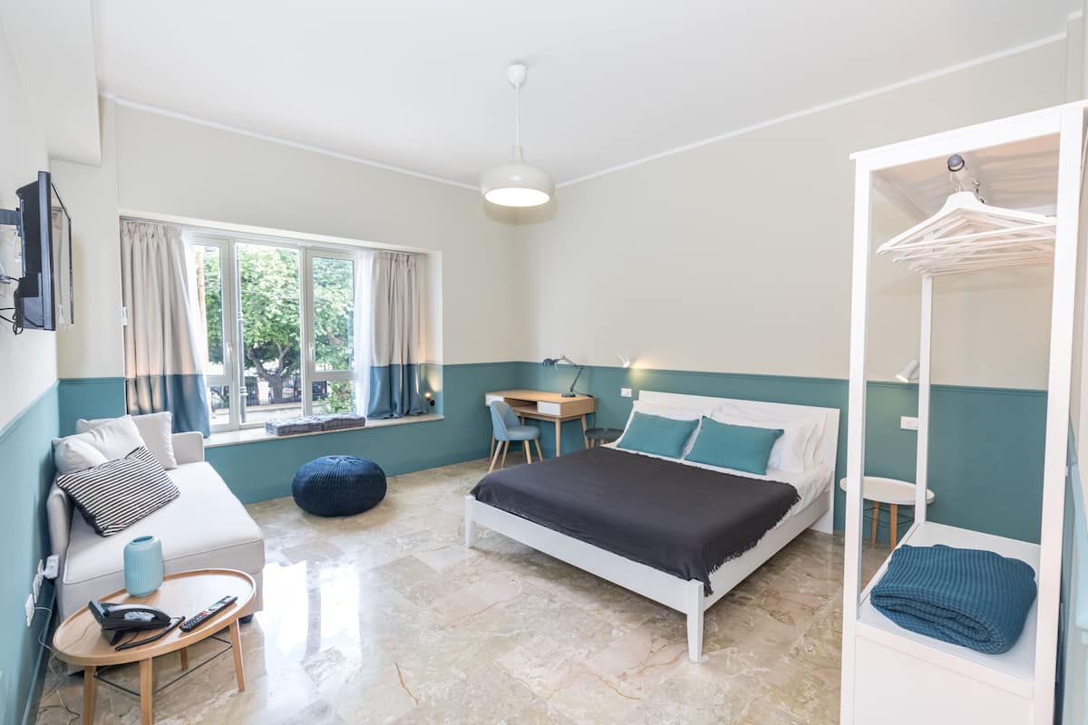A bedroom with a bed, a desk, a chair, a sofa, a hanging clothes rack, and a window with curtains.