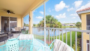 A balcony with a ceiling fan, a glass table, and chairs. There is a view of a body of water and greenery in the background. - Luxury Waterfront Condo Close to Fort Myers Beach and Golf Courses! (Fort Myers)