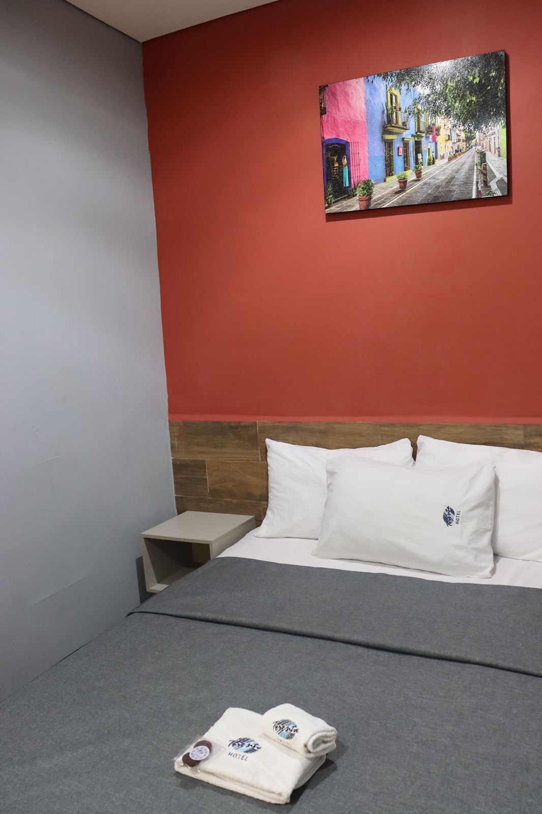 Una habitación de hotel con una cama, almohadas, una mesita de noche y un cuadro en la pared.