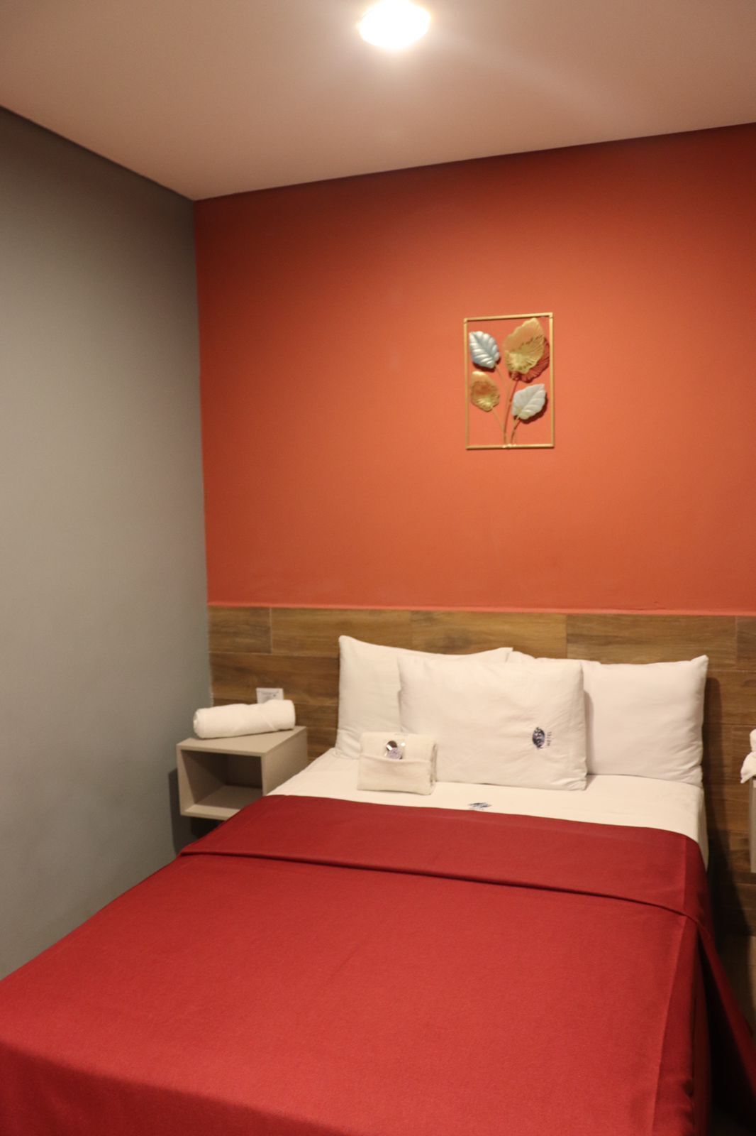A hotel room with a red bedspread, a wooden headboard, and a wall-mounted artwork of leaves.