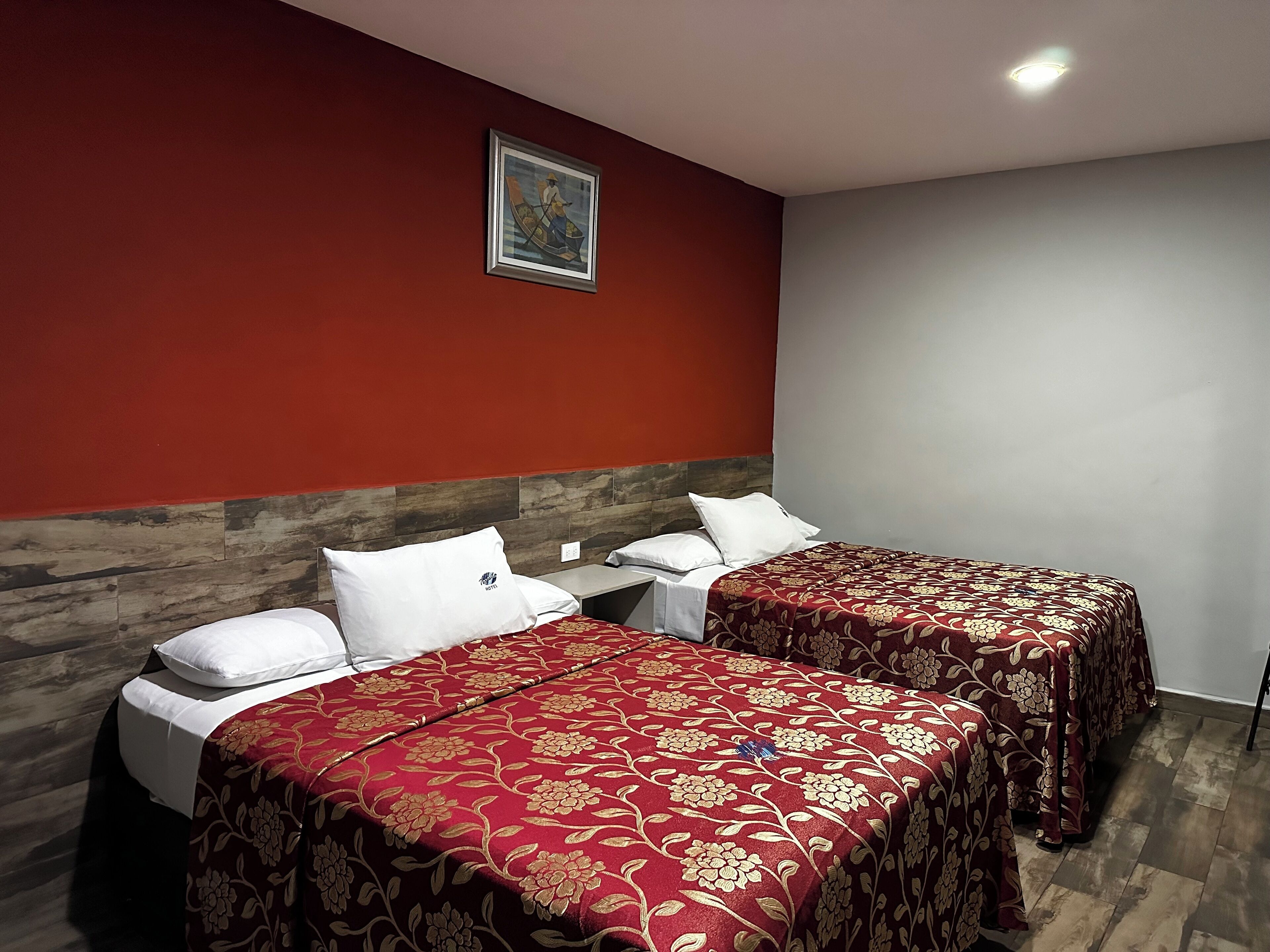 A hotel room with two beds, each with a red floral bedspread and white pillows. There is a wooden headboard, a small table with a phone, and a painting on the wall.