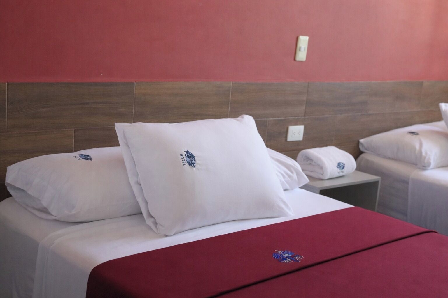 Habitación de hotel con dos camas, cada una con ropa blanca de cama y almohadas, un corredor de cama rojo como detalle y un cabecero de madera.