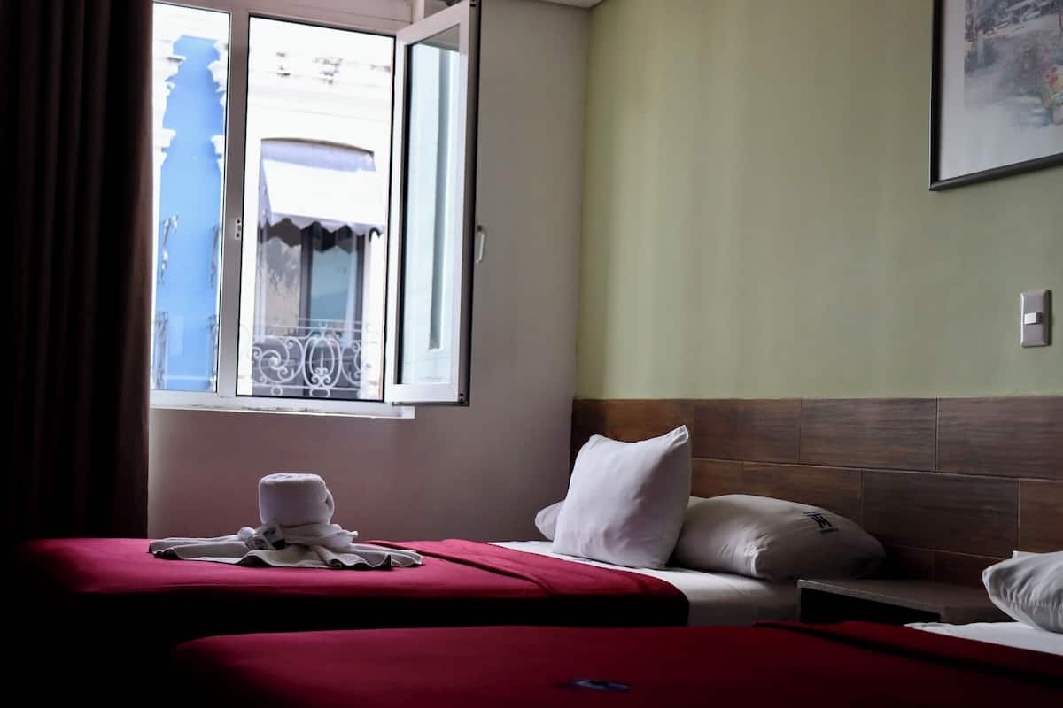 A hotel room with two beds, a window with a view of a building, and a painting on the wall.