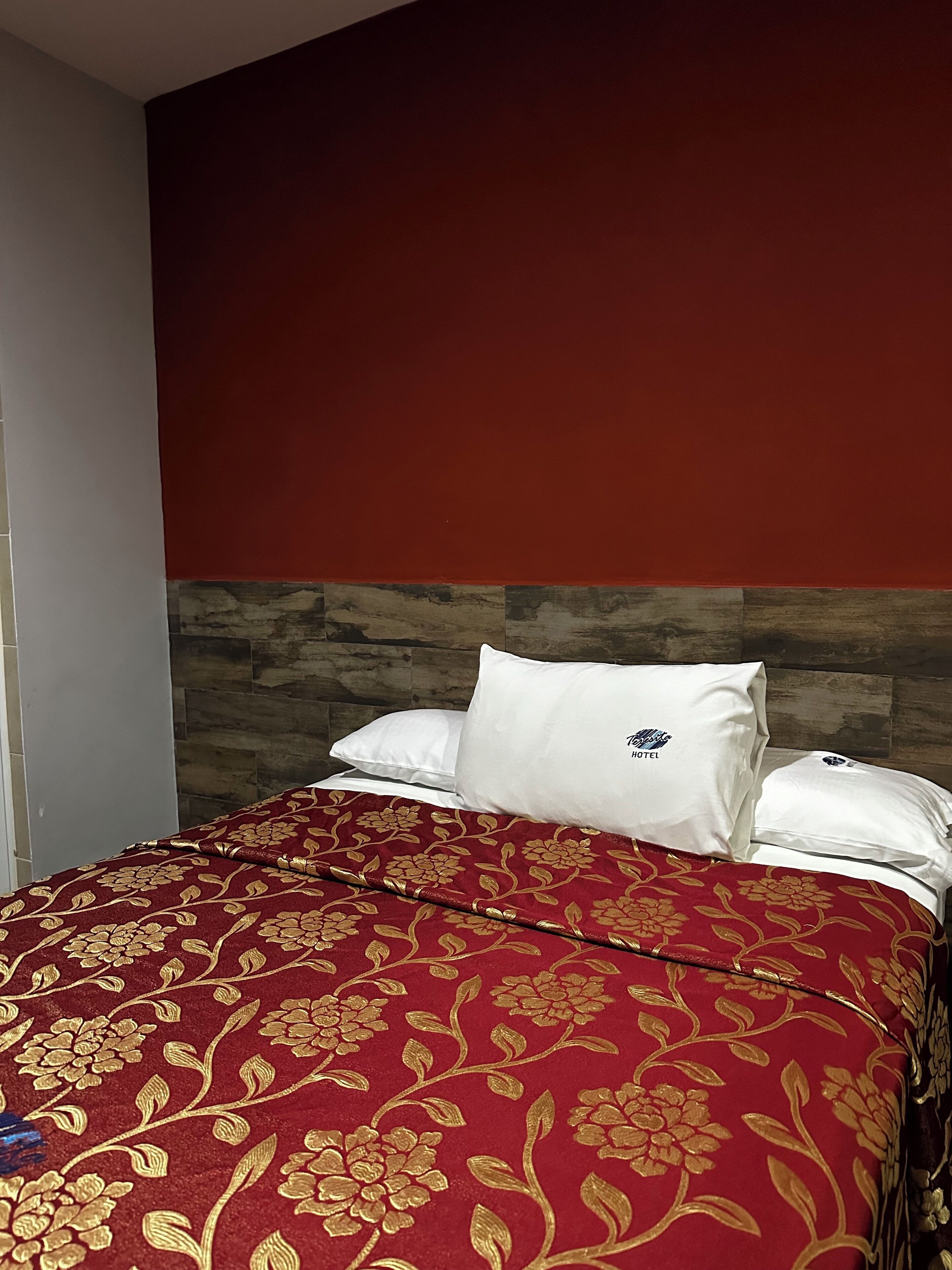 A hotel room with a bed, red and gold floral bedspread, white pillows, and a wooden headboard.