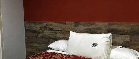 A hotel room with a bed, red and gold floral bedspread, white pillows, and a wooden headboard.