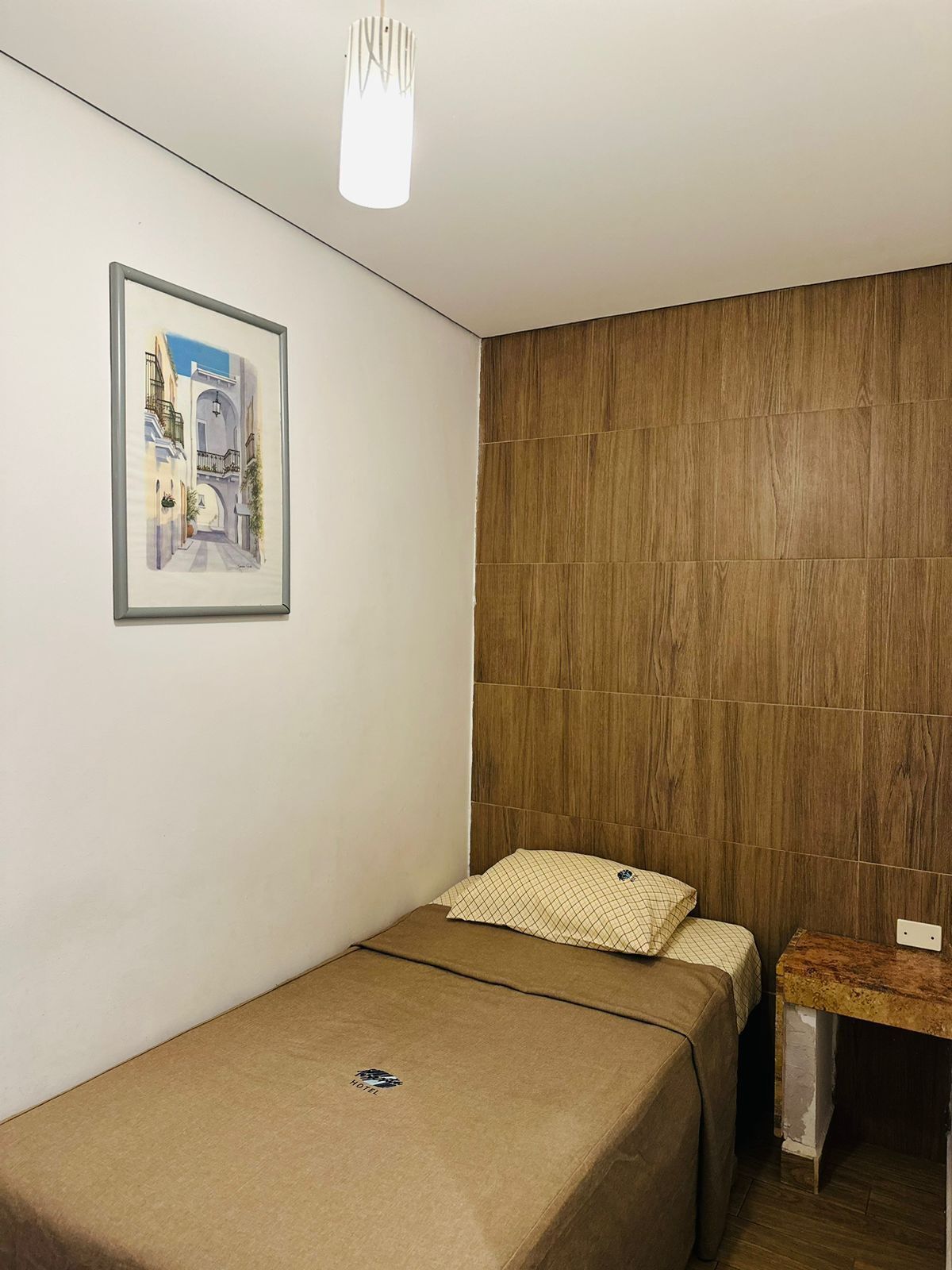A small, single-bed room with a wooden accent wall, a framed picture of a street scene, and a simple bedside table.