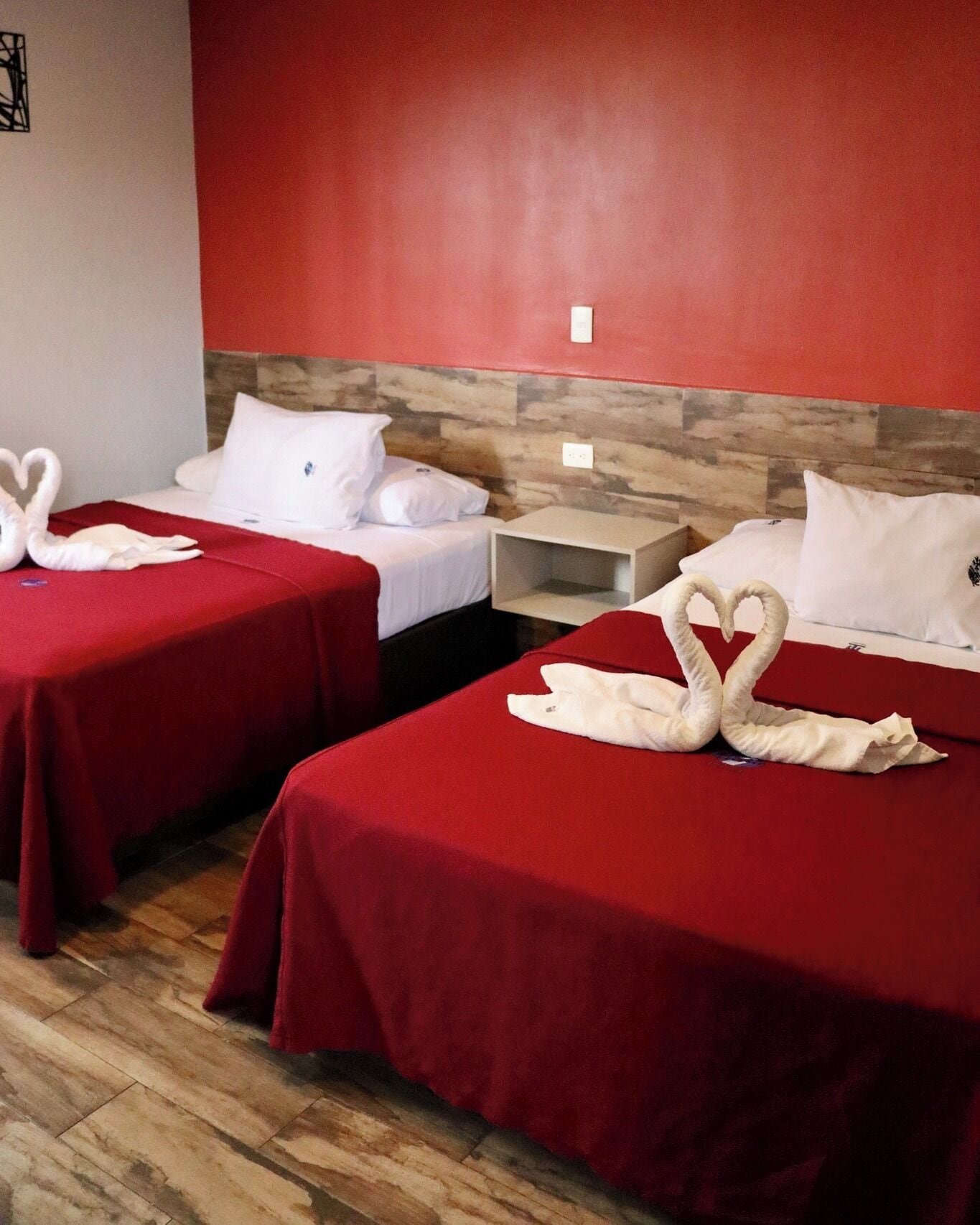 Two beds with red bedsheets and white pillows, each adorned with a swan-shaped towel. The room has a wooden headboard and a red accent wall.