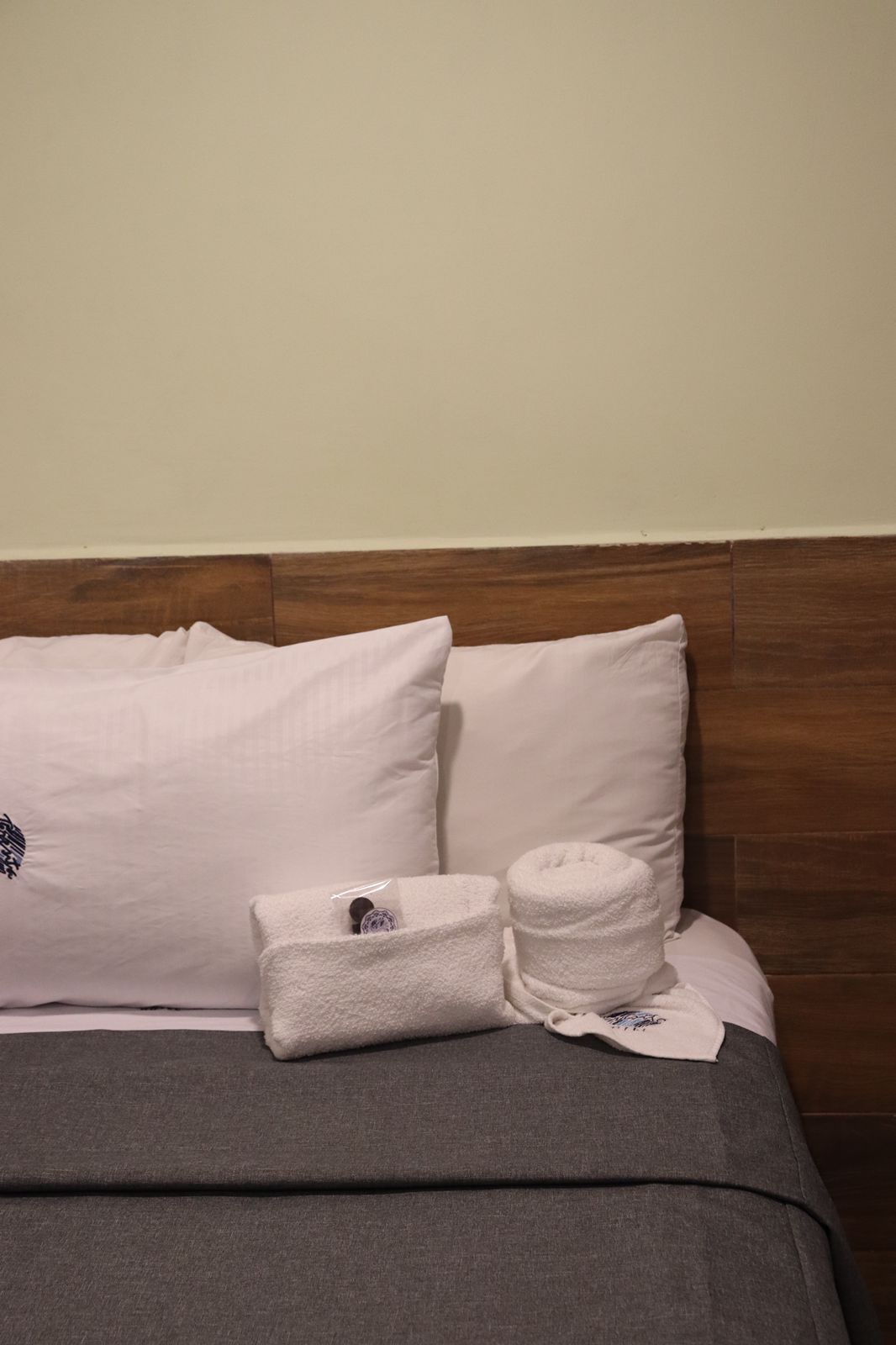 Ein ordentlich bezogenes Bett mit einer grauen Tagesdecke, zwei weißen Kissen und einem weißen Handtuch mit einem Logo.