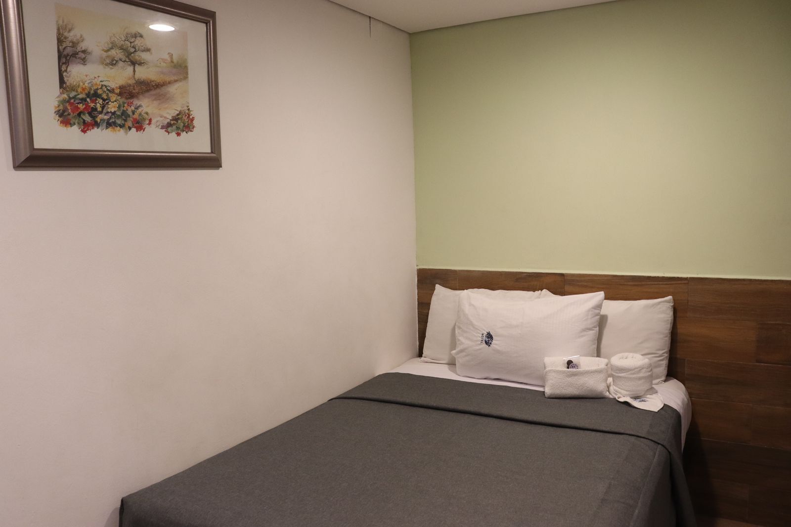 A single bed with a wooden headboard, white pillows, and a gray bedspread in a room with a light green wall and a framed painting above the bed.