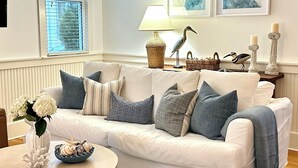 A living room with a white sofa, a round coffee table, and a lamp. There are two framed artworks on the wall, a window with blinds, and a bird figurine on the sofa. - Happy Hour Cottage Steps Away from Beach (Panama City Beach)