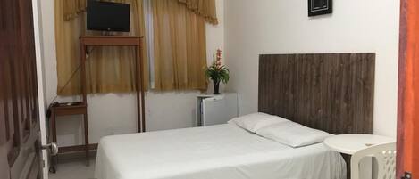 A hotel room with a bed, a small table, a chair, a TV, an air conditioner, a plant, and a framed picture on the wall.