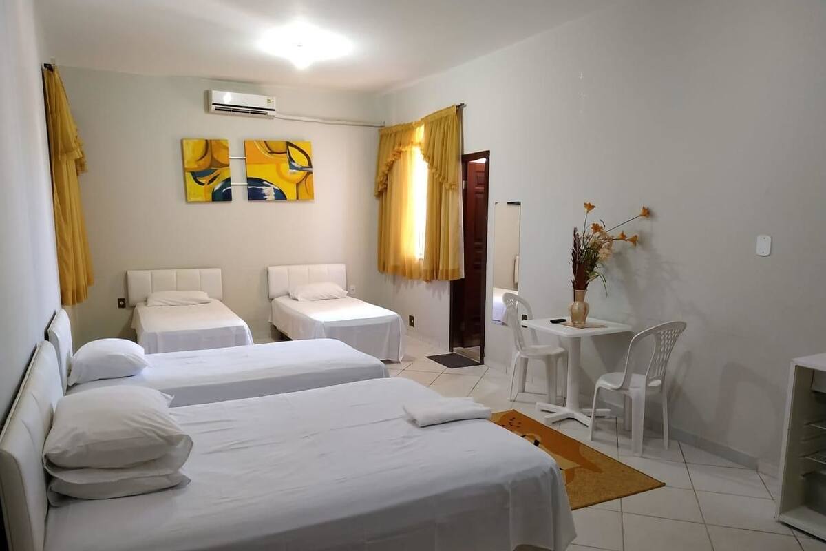Quarto de hotel com duas camas de solteiro, uma mesinha com cadeiras, um vaso com flores e um quadro na parede.
