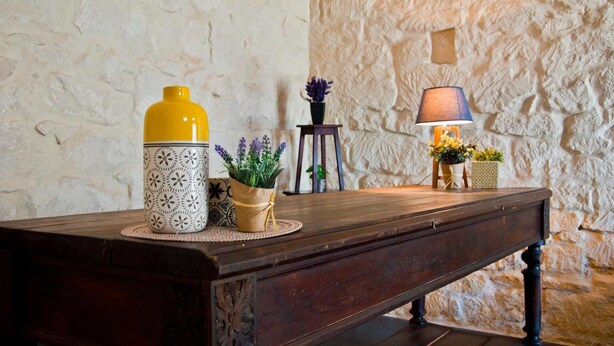 Una mesa de madera con un jarrón amarillo y blanco, una pequeña planta en maceta y una lámpara sobre un fondo de pared de piedra.