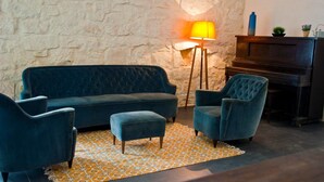 Una sala de estar acogedora con un sofá azul oscuro, sillones a juego y un pequeño reposapiés. Una alfombra estampada cubre el suelo, y una lámpara proporciona una iluminación cálida. Un piano está colocado contra la pared, y objetos decorativos se encuentran en un estante superior.