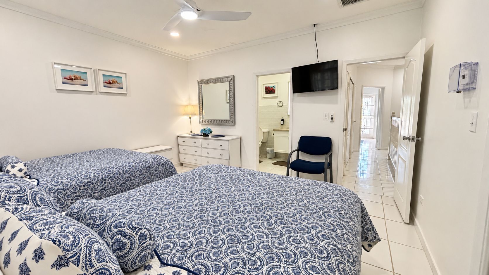 A bedroom with two beds, a dresser, a chair, a TV, and a bathroom visible through an open door.