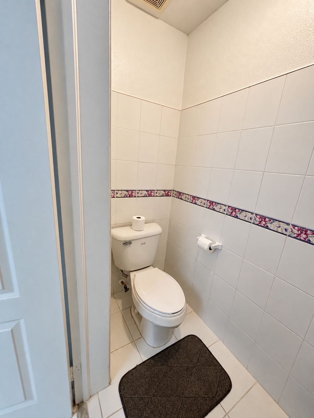A small, white bathroom with a toilet, a roll of toilet paper on the wall, and a brown bath mat on the floor.
