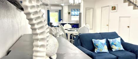 A modern living room with a grey sofa, two patterned cushions, a white decorative piece, and a dining area in the background.