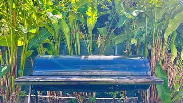 Un barbecue noir avec une table en métal, situé dans un jardin où poussent de grandes plantes vertes.