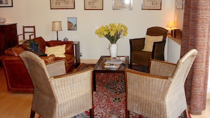 Una sala de estar con un sofá de cuero marrón, dos sillones de mimbre, una mesa de centro y un jarrón con flores amarillas. Hay cuadros en la pared y una lámpara en la mesita de noche.