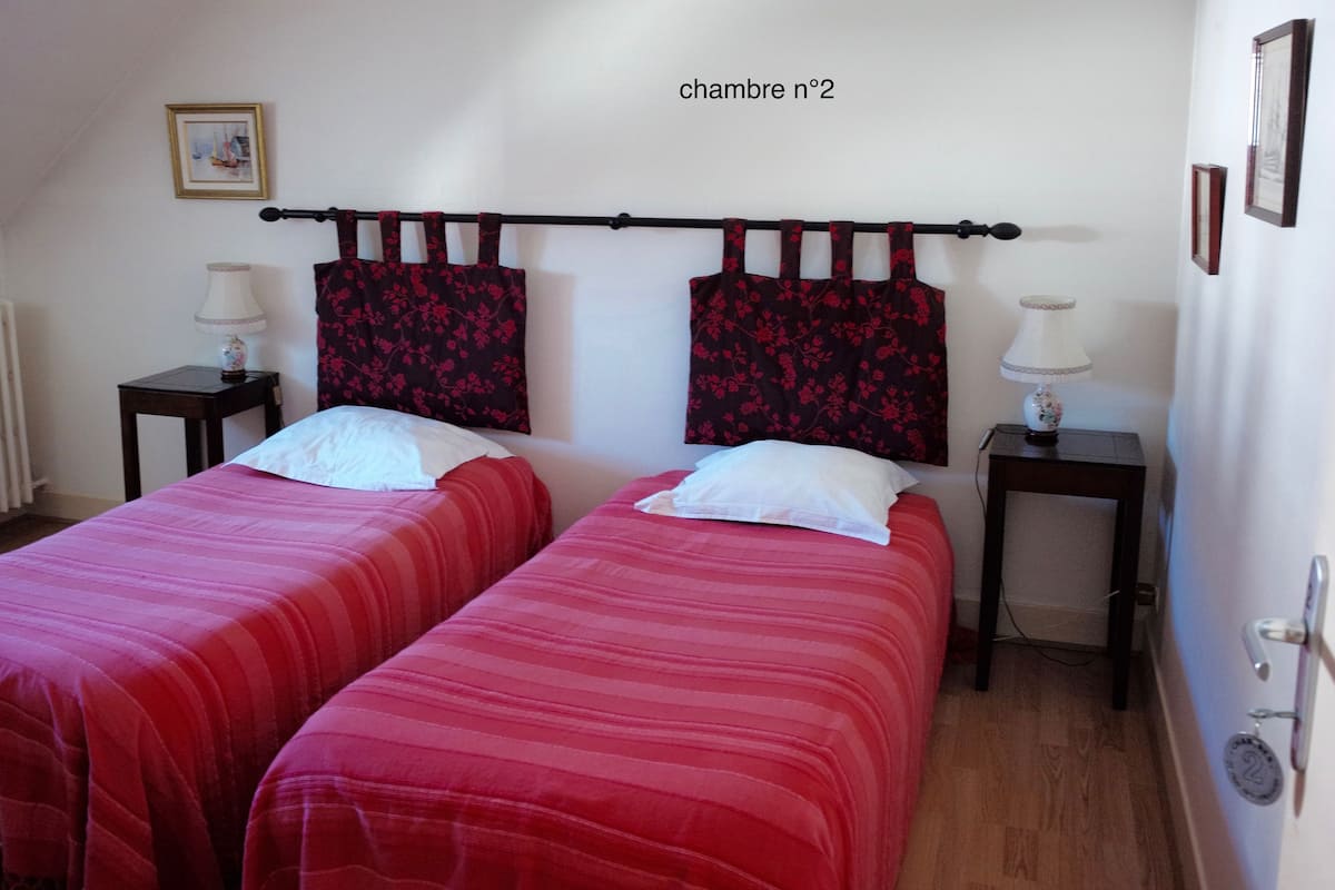 A room with two beds, each with a red striped bedspread and white pillows. There are two bedside tables with lamps, and a framed picture on the wall.