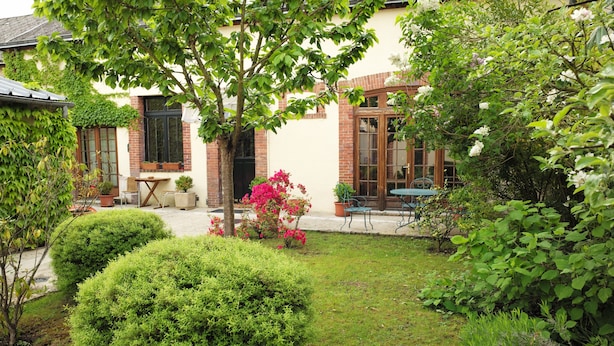 Un jardín bien cuidado con setos ordenados, una pequeña mesa y sillas, y un edificio con fachada de ladrillo y ventanas grandes.