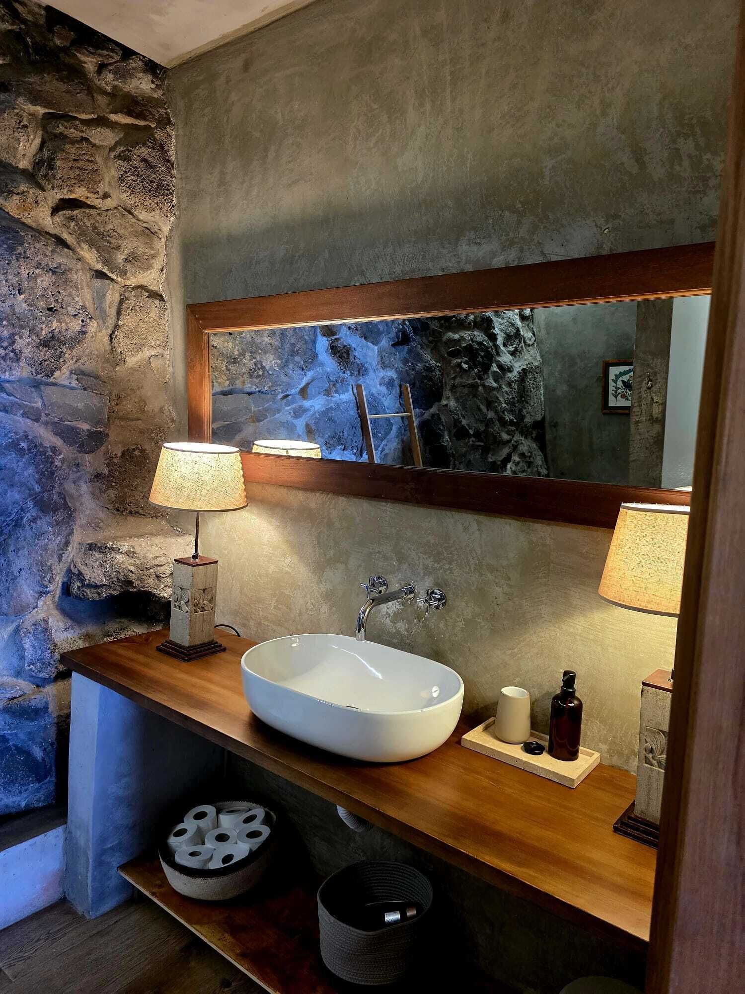 A bathroom with a stone wall, a large mirror, a white sink, and two lamps on a wooden countertop.