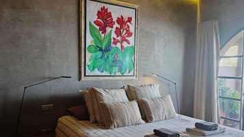 A bedroom with a bed, pillows, a bedside table, a lamp, and a painting of flowers on the wall.
