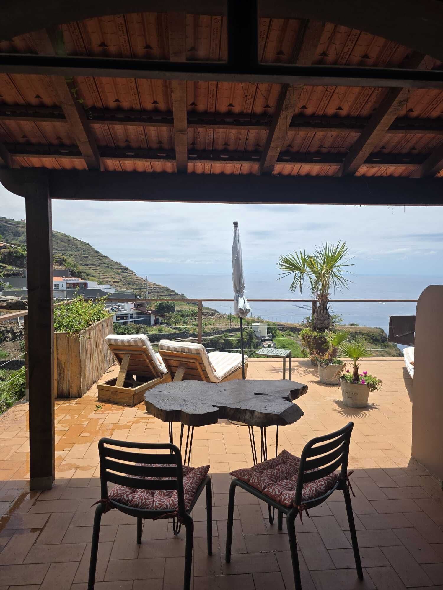 Un área de estar al aire libre techada con mesa y sillas, con vistas panorámicas al mar y las colinas.