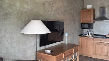 A compact living space with a wooden desk, a chair, a television, a kitchen area with cabinets and a microwave, and a concrete wall.