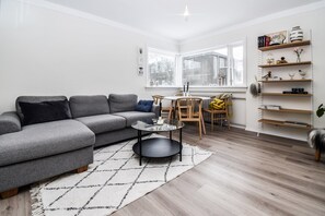 A modern living room with a grey sectional sofa, a glass coffee table, a dining area with a wooden table and chairs, and a white bookshelf with various items. - Apartment V20 (Reykjavik)