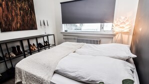 A bedroom with a bed, a radiator, a window with a blind, a shoe rack, and a floor lamp. - Apartment V20 (Reykjavik)