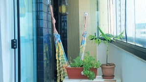 A balcony with a potted plant, a hanging macrame decoration, and a window with a view of buildings. - Duplex Caminho das Árvores (Salvador)