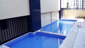 A rectangular indoor swimming pool with blue tiles, surrounded by a tiled deck and a white wall with a black vertical panel. - Duplex Caminho das Árvores (Salvador)