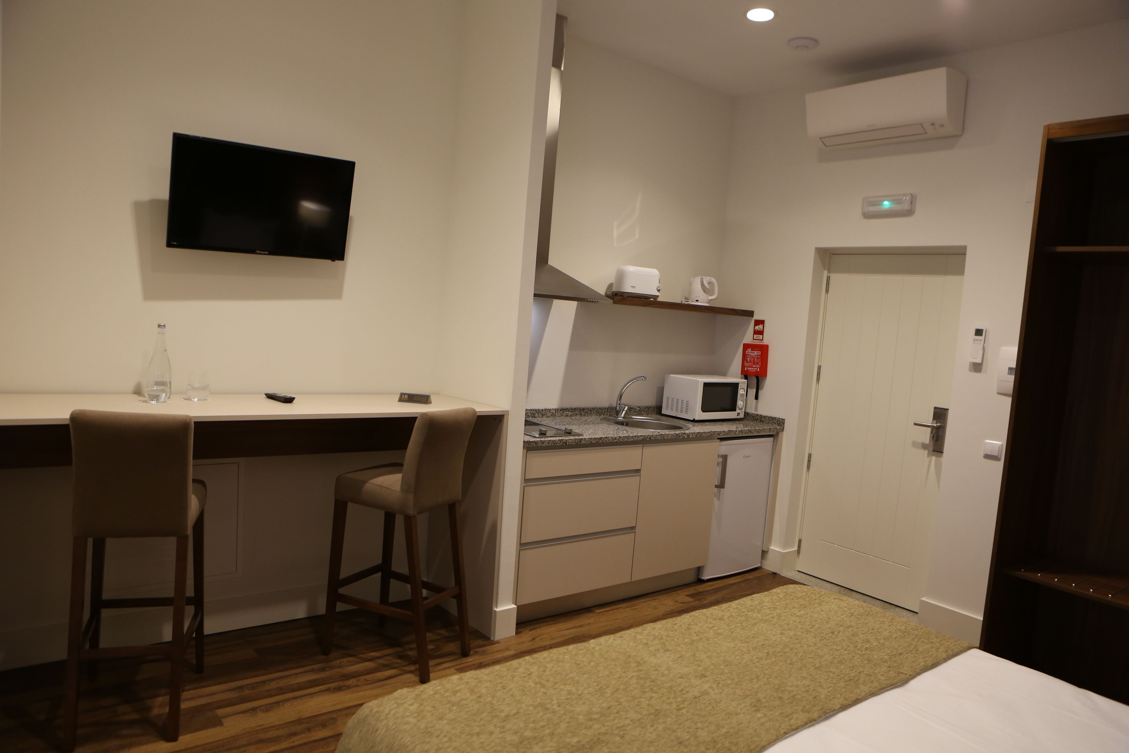 A compact hotel room with a kitchenette, a TV mounted on the wall, a bed, and a small seating area with two chairs.