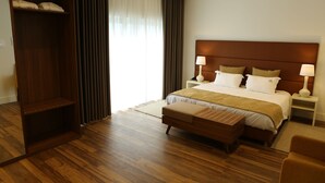 A modern hotel room with a wooden floor, a large bed with a headboard, two bedside tables with lamps, a wardrobe, and a large window with curtains. - Hotel Solar do Rebolo (Oliveira do Hospital)