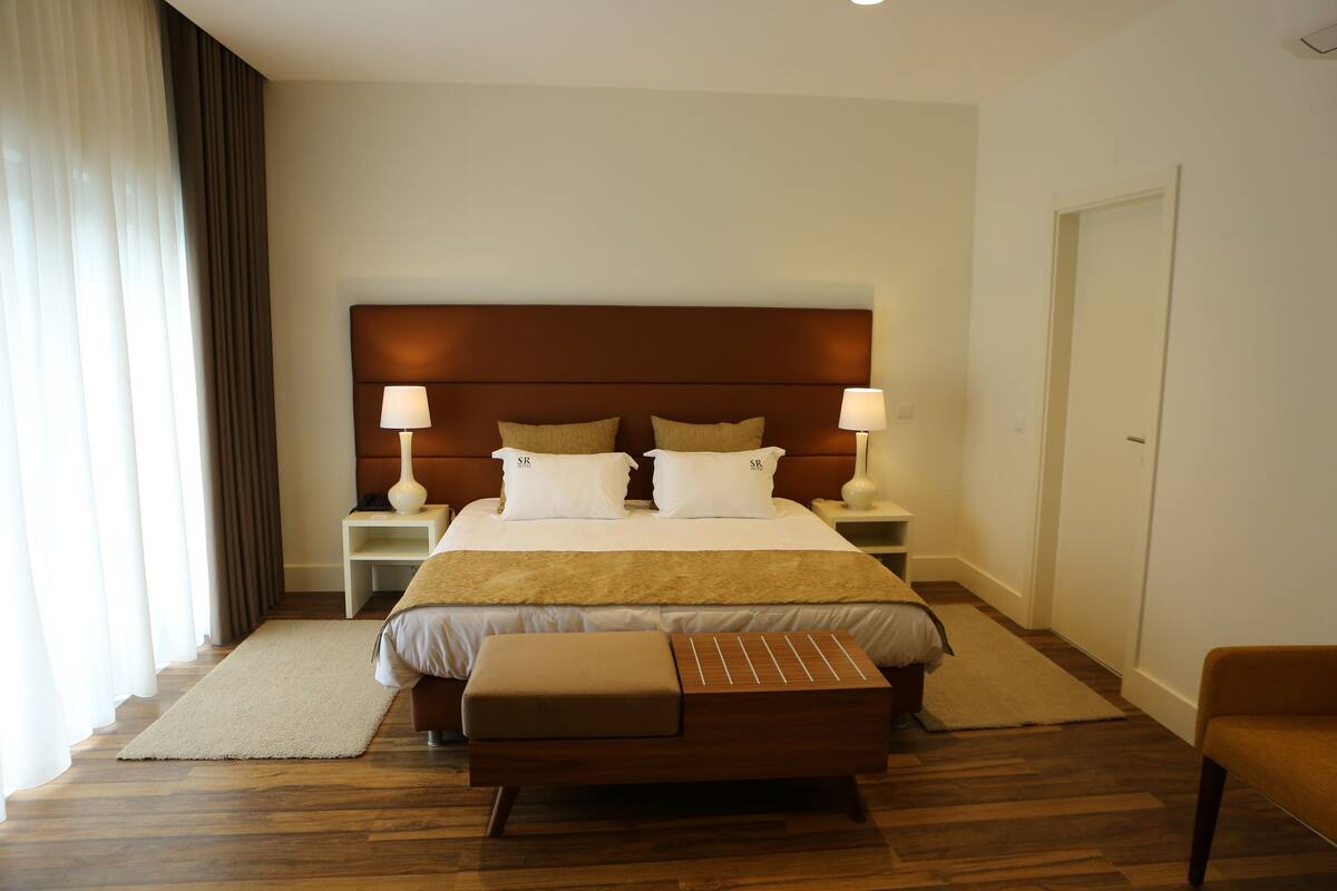 A hotel room with a large bed, two bedside tables with lamps, a wooden bench, and a view of the outside through a window with curtains.