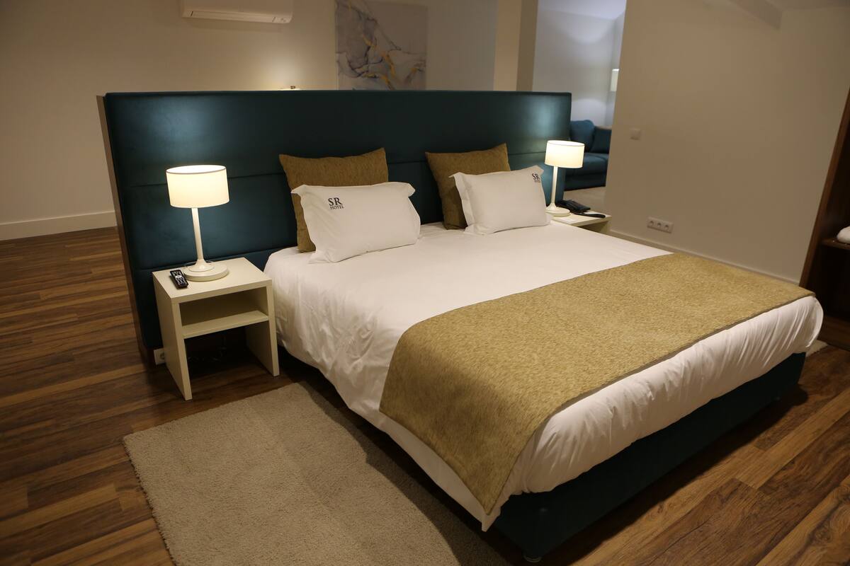 A neatly made bed with a dark headboard, white bedding, and two pillows with a logo. There is a bedside table with a lamp and a remote control. The room has wooden flooring and a beige rug.